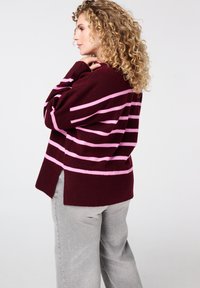 Bordeauxrode trui met roze strepen, met een relaxte pasvorm, zijsplitsingen en lange mouwen. Gecombineerd met lichtgrijze broek.