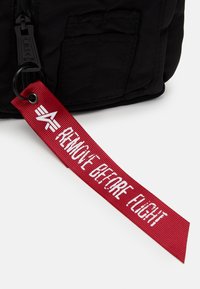 Černá nylonová taška s texturovaným povrchem, která má červenou tkanou etiketu s nápisem "ODSTRAŇTE PŘED LETEM" v bílém písmu.