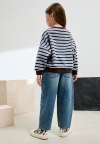 Suéter a rayas azul y marrón, de ajuste holgado, combinado con jeans anchos azul claro y zapatillas blancas con estampado. Fondo minimalista.