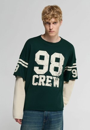 Giovane uomo con capelli biondi ricci indossa una camicia oversize verde scuro con stampa bianca "98 CREW" sopra una maglia beige a maniche lunghe e jeans blu.