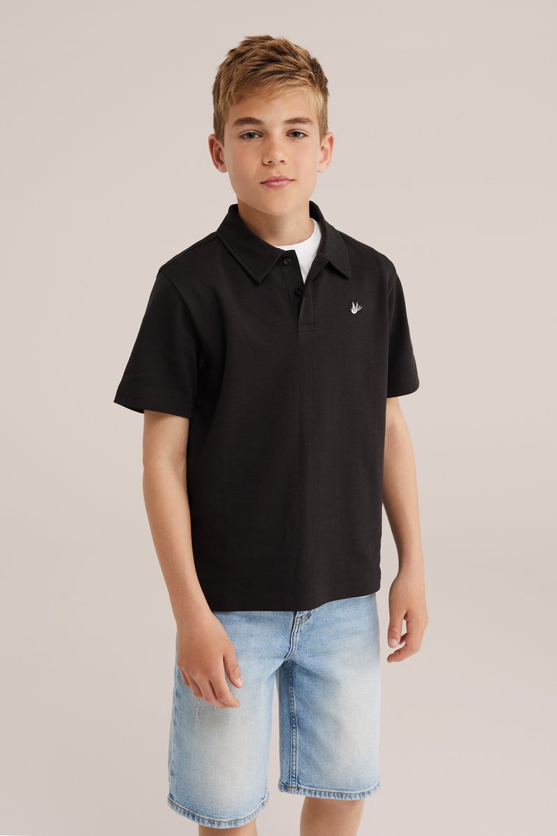 Garçon aux cheveux châtain clair portant un polo noir à manches courtes et un short en jean bleu clair, debout devant un fond beige uni.
