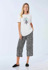 Weißes kurzärmliges T-Shirt mit Grafikdruck, kombiniert mit schwarzhäutigen, gemusterten weiten Hosen. Schwarze Slippers.