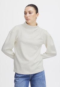 Haut crème à manches longues avec un col montant et des manches bouffantes, en tissu lisse et léger. Ourlet droit et coupe décontractée.