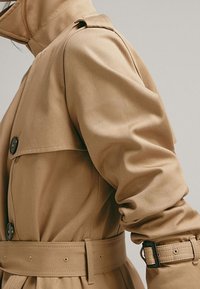 Tan trench coat with a structured collar, button details, and a belted waist. Made from a smooth, slightly textured fabric.