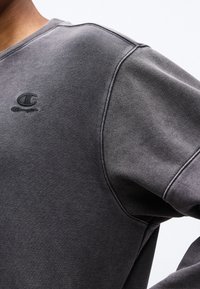 Κοντινό πλάνο ενός ατόμου που φοράει ξεβαμμένο μαύρο φούτερ Champion με κεντημένο λογότυπο στο στήθος, σε απλό φόντο.