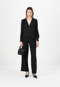 Femme se tenant debout dans une blouse noire à boutons, un pantalon à jambes larges, tenant un sac à main noir texturé, portant des escarpins à talons noirs sur fond blanc.
