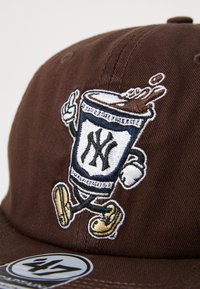 Brun hatt med brodert figur som holder en drink, med en blå og hvit logo, sammen med lyse sko og detaljerte sømmer.