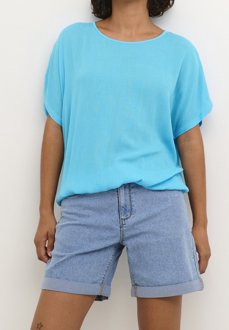 Femme portant un haut ample à manches courtes bleu clair et un short en jean retroussé, debout devant un fond blanc.