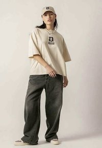 Young person wearing beige oversized t-shirt with graphic, wide dark jeans, beige cap, pearl necklace, and sneakers standing against plain background.