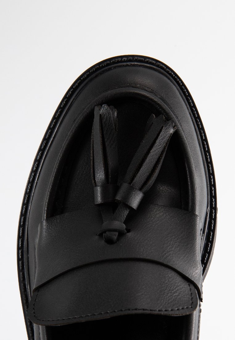 Mocassino in pelle nera con finitura liscia, dotato di due nappine e tacco piatto. Il design include cuciture attorno alla suola.