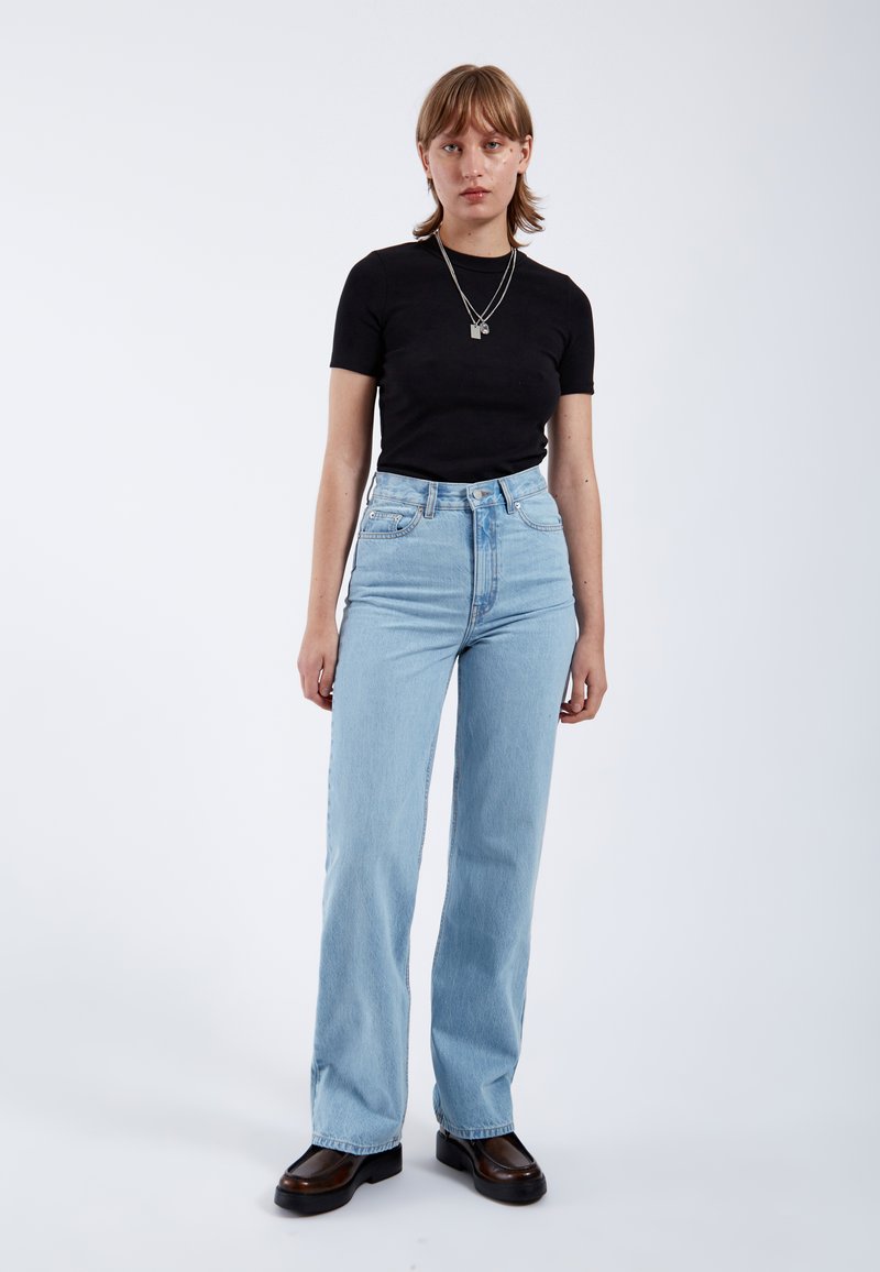 Jonge vrouw staat en draagt een zwart topje met korte mouwen, lichtblauwe hoge taille jeans, zwarte schoenen en gelaagde zilveren kettingen.