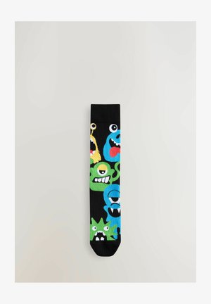 Chaussettes noires ornées de motifs de monstres colorés en vert, bleu et jaune avec des yeux et bouches expressifs, fabriquées à partir d'un matériau doux et flexible.