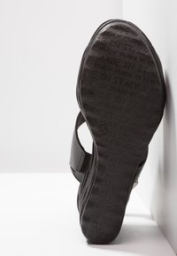 Semelle de sandale noire affichant en répétition le texte « Fabriqué en Italie » et la taille 37, avec une bride partiellement visible sur une surface blanche.