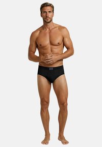 Slip in cotone nero con un design strutturato e una patch con il logo sulla fascia della vita, indossato da una figura maschile in piedi a piedi nudi.