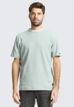 Man met kort bruin haar en baard, gekleed in een lichtgroen T-shirt met korte mouwen en een donkere spijkerbroek, staand tegen een effen lichte achtergrond.