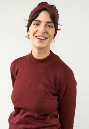 Bordeaux lange mouwtrui met een hoge hals. Het model draagt een bijpassende bordeaux hoofdband, wat eenvoud combineert met een gestructureerde afwerking.