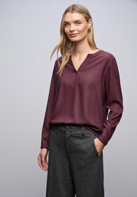 Bordeauxfarbene Bluse mit V-Ausschnitt, langen Ärmeln und glatter Textur, kombiniert mit grauen, hochgeschnittenen Hosen mit einem geknöpften Bund.