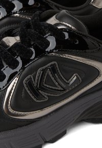 Melni sporta apavi ar lakota ādas akcentiem, teksturētām melnām auklām, izteiktu logotipu un masīvu gumijas zoli ar rievojumu apakšā.