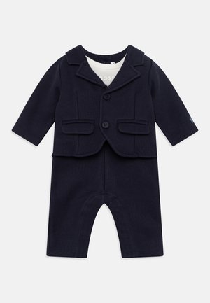Droit infantile bleu marine avec une veste style blazer, dotée de revers et de poches, accompagnée d'un pantalon long. Matière douce en tricot.