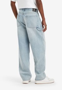 Világoskék denim farmernadrág, laza szabással, nagy hátsó zsebekkel és fekete, márkázott folttal a derékrészen. Egyszerű, klasszikus dizájn.