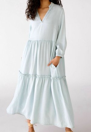 Femme portant une robe bleu clair à manches longues, col en V et à volants, avec des poignets élastiques et des poches latérales, debout avec une main dans la poche.
