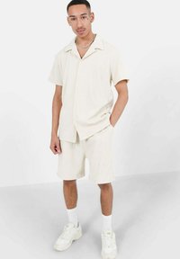 Camisa de manga corta de color crema y pantalones cortos a juego en tela texturizada. Presenta botones, un cuello y un corte relajado, combinados con zapatillas blancas.