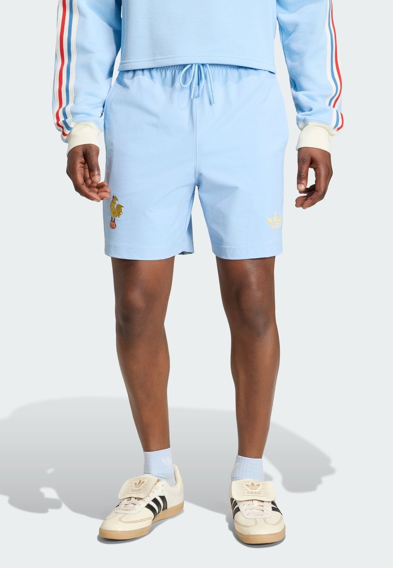 Hombre vistiendo pantalones cortos azul claro con logos dorados, sudadera a juego azul claro con mangas a rayas rojas, blancas y azules, y zapatillas Adidas blancas.