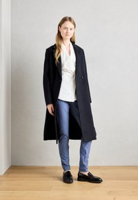 Navy vlněný kabát, střih na míru, do délky kolen. Bílá zavinovací blůza a světle modré slim fit kalhoty. Černé mokasíny dotvářejí vzhled.