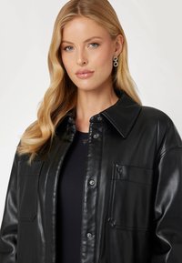 Giacca camicia in pelle nera con colletto, due tasche frontali e bottoni a pressione. Texture liscia e silhouette aderente.