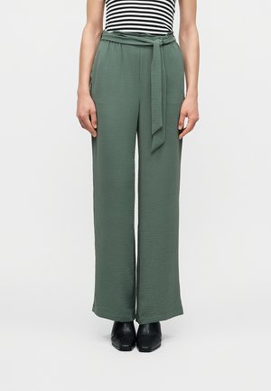 Γυναίκα που φοράει υφασμάτινο ζωνάκι στη μέση, πράσινο ψηλόμεσο παντελόνι φαρδύ και υφής, με μαύρα μυτερά παπούτσια.