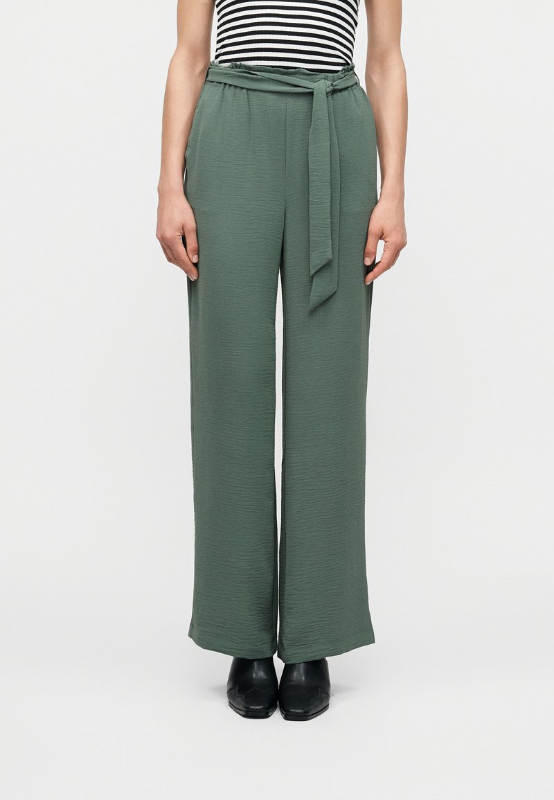 Γυναίκα που φοράει υφασμάτινο ζωνάκι στη μέση, πράσινο ψηλόμεσο παντελόνι φαρδύ και υφής, με μαύρα μυτερά παπούτσια.