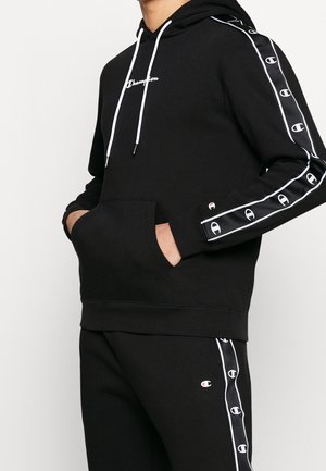 Mężczyzna w czarnej bluzie Champion z kapturem i dopasowanych spodniach dresowych z białymi paskami z logo na rękawach i nogawkach, ręce w kieszeni bluzy.