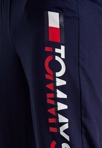 Marineblauwe sportbroek met een zachte textuur, voorzien van een gedurfde grafische uitstraling in rood, wit en zwart aan de zijkant met de tekst "TOMMY."
