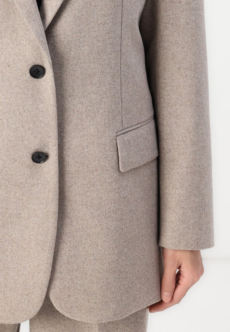 Blazer in lana marrone chiaro con design sartoriale, revers a punta, due bottoni neri e una tasca laterale. La texture è liscia e morbida.