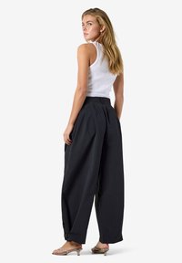 Femme debout, regardant par-dessus son épaule, vêtue d'un haut blanc sans manches, d'un pantalon noir ample et de sandales à talons, sur un fond uni.