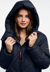 Marineblauwe gewatteerde pufferjack met een grote capuchon, ritsluiting en logodetail op de mouw. Zachte fleecevoering rond de capuchon.