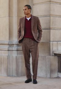 Brown checkered suit with a tailored fit, worn over a maroon knit shirt. Black shoes complete the outfit. Standing against stone backdrop.