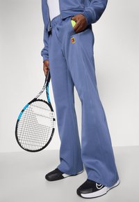 Ljusblå träningsoveraller med elastisk midja, som har en logotypapplikation och en sidoficka som håller en tennisboll, tillsammans med svarta skor.