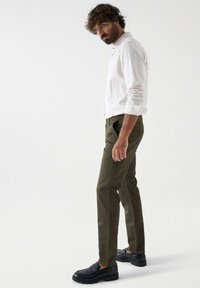 Olijf groene broek met een rechte snit en zijzakken, gecombineerd met een witte blouse met knopen en zwarte instappers, staand op een effen achtergrond.