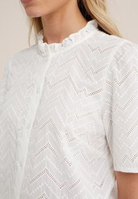 Vit skjorta med rynkad krage, prydd med ett zick-zack-mönster och ögonlettsdetaljer. Korta ärmar i ett texturerat, lätt material.