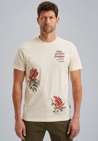 T-shirt en coton couleur crème avec des impressions graphiques rouges et vertes et du texte sur le devant. Manches courtes, col rond, coupe décontractée.
