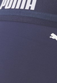 Temno modri atletski leggingi z širokim pasom, na katerem je bela blagovna znamka "PUMA", ter majhnim belim logotipom na spodnji strani. Gladka tekstura tkanine.