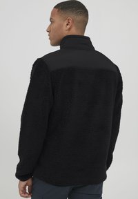 Schwarze Fleecejacke mit hohem Kragen und einer glatten Stoffeinlage. Verfügt über ein strukturiertes, flauschiges Material an Körper und Ärmeln.