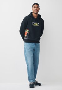 Zwarte hoodie met grafische print van cartoonfiguren, kangaroo-zak, capuchon met trekkoord, gecombineerd met lichtblauwe jeans en zwarte sportschoenen.