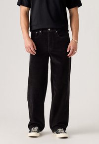 Personne portant un pantalon en velours côtelé noir ample, un t-shirt noir, des baskets noir et blanc, se tenant devant un fond clair uniforme.