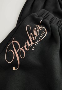 Černá látka s hladkou texturou, na které se nachází výrazné růžově zlaté kurzívou napsané logo "Baker" a text "by Ted Baker" pod ním.