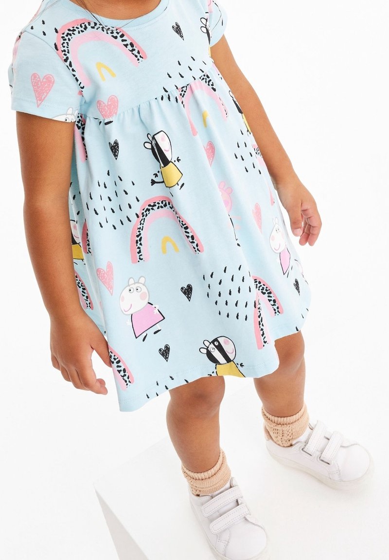 Tout-petit portant une robe bleu clair avec des personnages de dessin animé, des arcs-en-ciel, des cœurs, des chaussettes beiges et des chaussures blanches à scratch, debout sur une surface blanche.