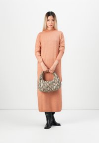 Egy nő áll, őszibarack színű, hosszú pulóver ruhát visel, fekete térd fölé érő csizmát, és egy kígyóbőr mintájú táskával áll a fehér háttér előtt.
