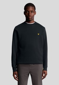 Zwarte sweatshirt met ronde hals en lange mouwen, met een klein geel logo op de linkerborst. Draagt met grijze broek.