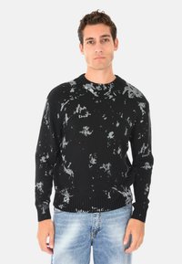 Maglione nero con motivi astratti grigi, polsini e orlo a coste. Indossato sopra jeans strappati blu chiaro. Girocollo.